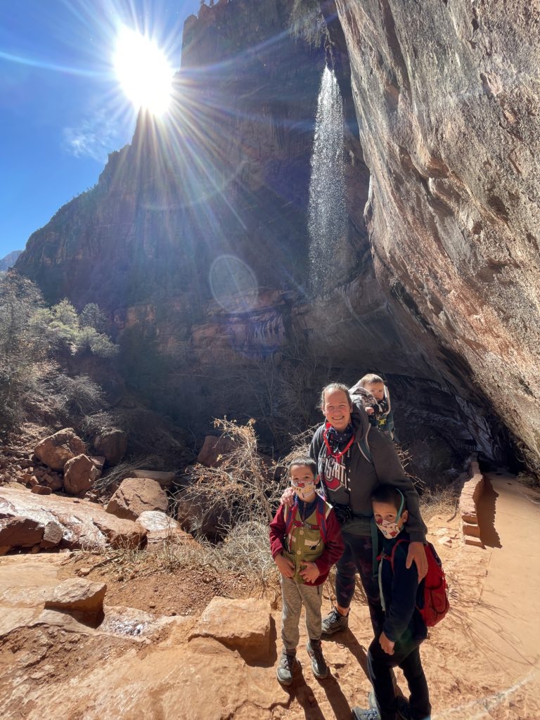 A woman and her three children stand under one of the waterfalls in the Lower Emerald Pool hike at Zion National Park. - Exploring Through Life