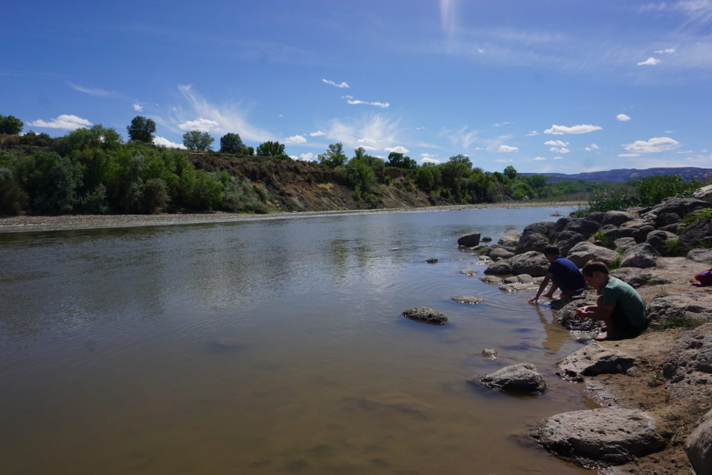 Two young children play at the edge of the Colorado River at Las Colonias Park. The river is wide and calm. A great family-friendly attraction in Grand Junction.