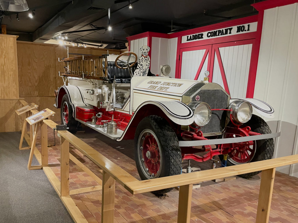 A vintage fire truck with a stuffed Dalmatian dog in the drivers seat. The wall behind the truck says Ladder Company No. 1. The truck is at the Museum of the West.